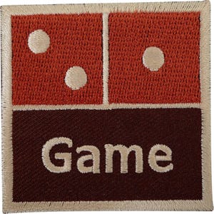May include: A square embroidered patch featuring a rust-orange top section with three cream-coloured dots arranged like a domino, and a dark brown bottom section with the word Game stitched in cream-coloured thread, all bordered by a cream edge.