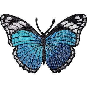 Puede incluir: Un parche bordado de mariposa con alas de color azul vibrante y azul marino oscuro, con detalles en blanco a lo largo de los bordes superiores. La mariposa tiene un cuerpo central negro y dos antenas negras finas hacia arriba.