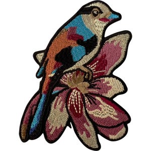 May include: An embroidered patch featuring a bird with blue, orange, and black plumage perched atop a large, multi-petaled flower in shades of pink, burgundy, and cream. The design is outlined in a thick black border, creating a distinct shape.