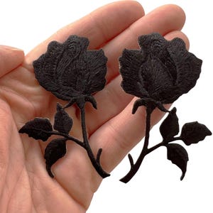 May include: Two black rose patches featuring detailed embroidery. Each patch depicts a single rose bloom with a stem and several leaves. The embroidery uses dense black thread to create texture and depth in the petals and foliage of the flowers.