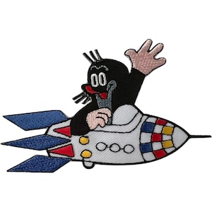 May include: An embroidered patch featuring a black mole character with a red nose and white eyes, waving a hand while piloting a white rocket ship. The rocket has blue fins, red and yellow accents, and three circular windows on its side.