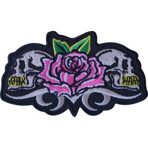 May include: An embroidered patch featuring a central pink rose with green leaves, flanked by two light gray skulls on a black background. The design includes curved light gray accents at the bottom, all outlined in black thread on a dark fabric base.