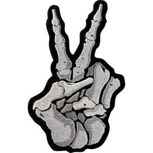 May include: An embroidered patch featuring a skeletal hand making a peace sign gesture. The bones are rendered in shades of light gray and white thread, with dark gray shading for depth, all outlined by a thick black border on a black background.