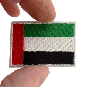 May include: A rectangular embroidered patch featuring the flag of the United Arab Emirates. The design consists of a vertical red stripe on the left side, followed by three horizontal stripes in green, white, and black, all bordered by white stitching.