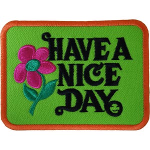May include: A rectangular embroidered patch with a bright lime green background and an orange border. It features a pink flower with a teal centre and green leaves, alongside the black embroidered text "HAVE A NICE DAY" with a smiley face.