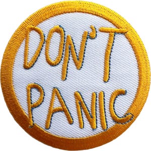 Puede incluir: Un parche bordado circular con fondo blanco y un borde grueso de color amarillo dorado. El texto DON'T PANIC está bordado con hilo amarillo dorado en el centro en una fuente de estilo manuscrito informal con contornos azul oscuro.