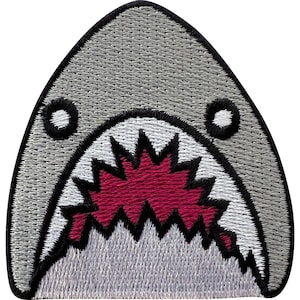 May include: An embroidered patch shaped like a shark head with a gray exterior, a white jaw area, and a dark red interior mouth featuring sharp, jagged black-outlined teeth. The entire design is bordered by a thick black stitched edge.