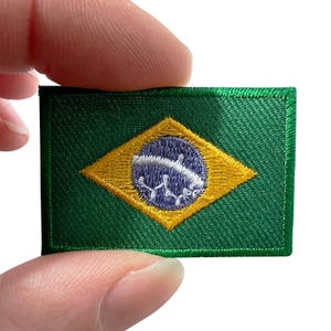 May include: A rectangular embroidered patch featuring the flag of Brazil. The design consists of a green field with a central yellow rhombus containing a blue circle. Inside the blue circle is a white curved band with small white star-like details.
