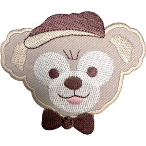May include: An embroidered patch featuring the head of a light brown teddy bear wearing a dark brown beret and a matching dark brown bow tie. The bear has large white circular patches around its dark brown eyes, a small nose, and a pink tongue.