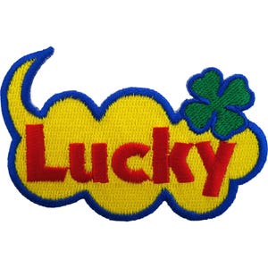 May include: An embroidered patch featuring the word Lucky in bold red letters centered on a yellow cloud-shaped background. The design is outlined in blue thread and includes a small green four-leaf clover positioned on the upper right side.