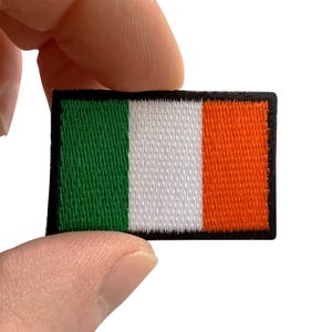 May include: A rectangular embroidered patch featuring the national flag of Ireland, consisting of three vertical stripes in green, white, and orange, with a solid black border surrounding the entire design on a white background.