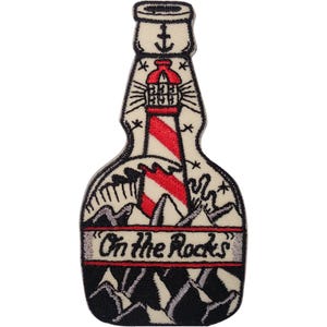 Könnte beinhalten: Ein gestickter Aufnäher in Flaschenform mit cremefarbenem Hintergrund. Er zeigt einen rot-weiß gestreiften Leuchtturm, schwarze Bergsilhouetten und den Text "On the Rocks" in schwarzer Schrift. Am Flaschenhals befindet sich ein kleiner schwarzer Anker.