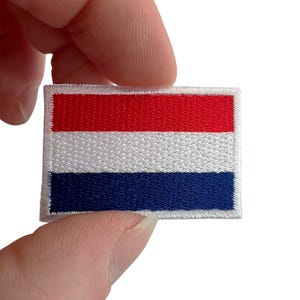May include: A rectangular embroidered patch featuring the horizontal tricolor flag of the Netherlands, consisting of three equal bands of red, white, and blue, with a white stitched border surrounding the entire edge of the fabric patch.