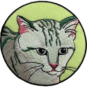 Op de afbeelding: Een ronde geborduurde patch met een witte kat met grijze tabby-markeringen en roze binnenoren. De kat is geplaatst tegen een licht limoengroene achtergrond en het ontwerp is omlijnd met een dikke, stevige zwarte geborduurde rand.