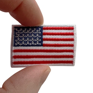 May include: A rectangular embroidered patch featuring the United States flag. The design includes a blue canton with white stitched stars and alternating horizontal red and white stripes. The patch has a white stitched border around the entire edge.