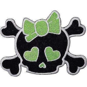 Puede incluir: Un parche bordado que presenta una calavera negra con huesos cruzados detrás. La calavera tiene dos ojos verdes brillantes en forma de corazón, una pequeña nariz triangular verde y un gran lazo verde en la parte superior, todo bordeado en blanco.
