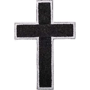 Puede incluir: Un parche bordado en forma de cruz negra que presenta una sección central negra gruesa rodeada por un borde blanco fino. El bordado tiene una apariencia texturizada y elevada con bordes claramente definidos que forman una cruz.