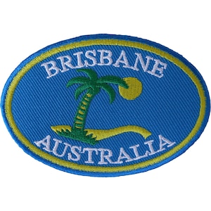 May include: An oval-shaped embroidered patch featuring a blue background with a yellow border. The text BRISBANE is stitched in white at the top, and AUSTRALIA is stitched in white at the bottom. A green palm tree and yellow sun are centered.