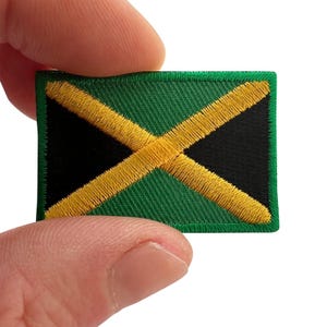 May include: A rectangular embroidered patch featuring the flag of Jamaica. The design consists of a gold saltire cross dividing the patch into four triangles, with two green triangles on the top and bottom and two black triangles on the left and right sides.
