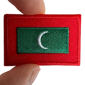May include: A rectangular embroidered patch featuring a red border surrounding a green center rectangle. In the middle of the green section is a white crescent moon symbol. The embroidery has a visible thread texture throughout the design.
