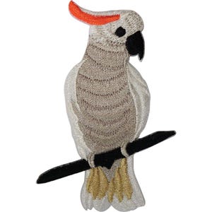 May include: An embroidered patch featuring a cockatoo with light grey and white plumage, a bright orange crest, and a black beak. The bird is perched on a horizontal black branch, and its tail feathers are a light golden yellow colour.