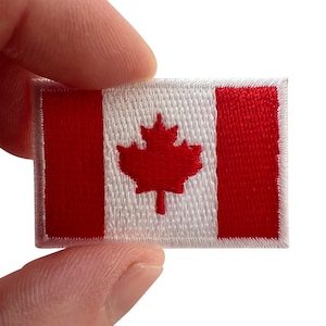 May include: A rectangular embroidered patch featuring the Canadian flag design. The patch displays a central red maple leaf on a white background, flanked by two vertical red bars on the left and right sides, all finished with a white stitched border.