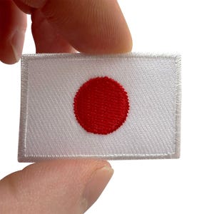 Puede incluir: Un parche bordado rectangular que presenta la bandera nacional de Japón. El diseño consiste en un fondo blanco sólido con un emblema circular rojo centrado. Los bordes del parche están acabados con un borde cosido de color blanco.