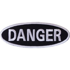 May include: An oval-shaped embroidered patch featuring a black background with the word DANGER stitched in bold white capital letters. The patch is finished with a thick white border that outlines the entire oval shape, creating a high-contrast design.