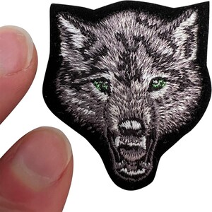 Peut inclure: Un écusson brodé représentant la tête d'un loup au pelage gris et blanc, avec des dents acérées et des yeux verts perçants. Le loup est représenté gueule ouverte avec une expression menaçante sur un fond de forme noire unie.
