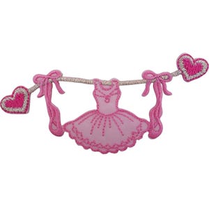 Peut inclure: Un écusson brodé représentant une robe de ballet rose clair centrée sur une ligne horizontale. La ligne est décorée de deux nœuds roses et de deux cœurs roses avec des bordures argentées aux extrémités. Le motif est bordé de fil rose.