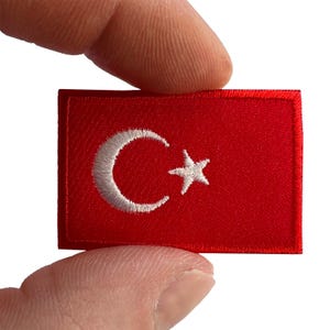 Puede incluir: Un parche bordado rectangular que presenta la bandera de Turquía. El diseño consiste en un fondo rojo sólido con una luna creciente blanca y una estrella blanca de cinco puntas centradas en el lado izquierdo del parche de tela con bordes cosidos.
