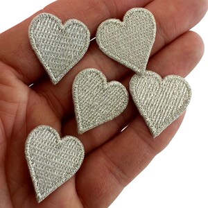May include: Five small heart-shaped patches featuring a metallic silver thread embroidery with a dense, textured stitch pattern and a finished border. Each heart has a consistent shape and reflective sheen, designed for application as decorative accents.