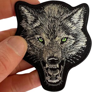 Pode incluir: Um emblema bordado apresentando a cabeça de um lobo com pelo cinza e branco, dentes afiados e olhos verde-brilhantes. O design é definido contra um fundo preto sólido com uma borda preta espessa, criando uma aparência detalhada.