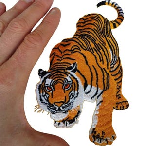 May include: An embroidered tiger patch featuring a vibrant orange coat with black stripes, white accents on the face and underbelly, and piercing red eyes. The tiger is depicted in a prowling stance with its tail curved upward and visible stitching.