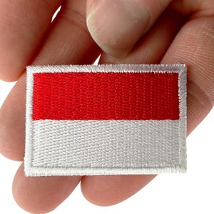 May include: A rectangular embroidered patch featuring a horizontal bicolor design with a solid red upper half and a solid white lower half, finished with a white stitched border around the entire perimeter of the fabric emblem.
