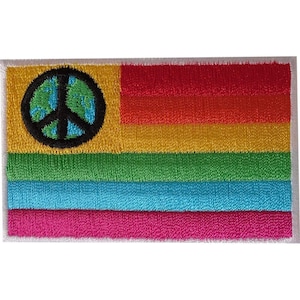 Op de afbeelding: Een rechthoekige geborduurde patch met een horizontaal streeppatroon in rood, oranje, geel, groen, blauw en magenta. Een zwart cirkelvormig vredessymbool met een blauwe en groene wereldbol bevindt zich aan de linkerkant.