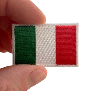 May include: A rectangular embroidered patch featuring the Italian flag design with vertical stripes of green, white, and red. The patch has a white stitched border around the entire perimeter, showcasing a dense thread texture throughout the design.