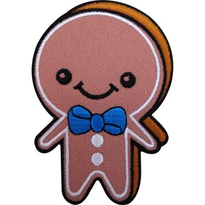 Puede incluir: Un parche bordado con forma de hombre de jengibre con piel de color marrón claro, una pajarita azul brillante y dos botones circulares blancos en el torso. La figura presenta una sonrisa negra bordada, ojos negros y un borde blanco.