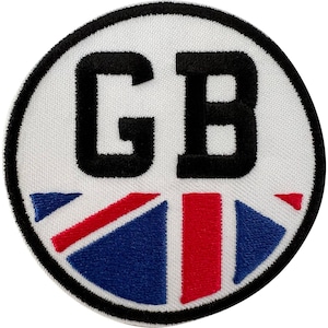 May include: A circular embroidered patch featuring a white background with the black letters GB centered at the top. The lower portion of the patch displays a stylized Union Jack flag design in red, white, and blue, all enclosed by a black border.