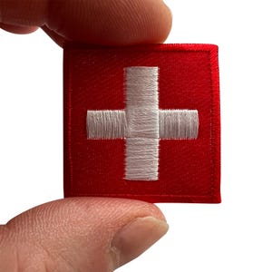 Op de afbeelding: Een vierkante geborduurde patch met een felrode achtergrond en een wit kruis in het midden. Het ontwerp is gestikt met een dicht draadpatroon, wat zorgt voor een getextureerd oppervlak dat de vlag van Zwitserland in een compact formaat nabootst.