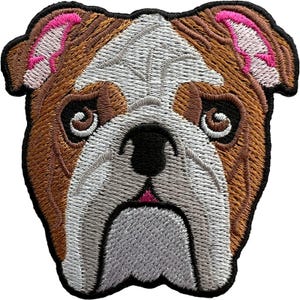 Può includere: Una toppa ricamata raffigurante la testa di un bulldog con pelo marrone e bianco. Il cane ha grandi occhi scuri, un naso nero e accenti rosa all'interno delle orecchie. La toppa è bordata con filo nero, che ne definisce la forma del muso.