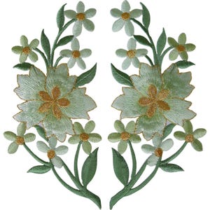Könnte beinhalten: Zwei symmetrische, gestickte Blumenaufnäher mit hellgrünen Blütenblättern und goldgelben Mitten. Jeder Aufnäher zeigt eine große zentrale Blüte, umgeben von kleineren Blüten und spitz zulaufenden grünen Blättern in detaillierter Stickerei.