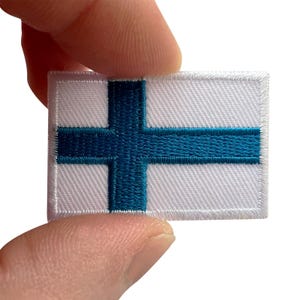 Puede incluir: Un parche rectangular bordado con la bandera de Finlandia, que consiste en un fondo blanco con un diseño de cruz nórdica en azul oscuro. El parche tiene un borde cosido en blanco y se sostiene entre dos dedos sobre un fondo blanco.