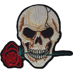 Op de afbeelding: Een geborduurde patch met een beige menselijke schedel met een zichtbare barst aan de linkerbovenkant. De schedel heeft donkere oogkassen en houdt een felrode roos met een groene steel en bladeren tussen de tanden, omlijnd met zwart garen.