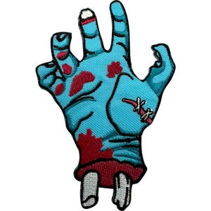 Peut inclure: Un écusson brodé en forme de main coupée avec une peau bleu clair, des éclaboussures de sang rouge et des fragments d'os blancs visibles au poignet. La main présente des contours noirs, une cicatrice cousue et une base rouge foncé.