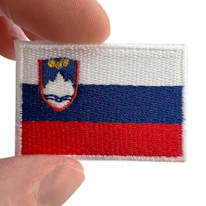 May include: A rectangular embroidered patch featuring the national flag of Slovenia. The design consists of three horizontal stripes in white, blue, and red, with the Slovenian coat of arms positioned on the upper left side, displaying a mountain.