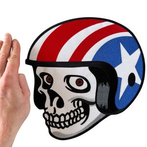May include: A large embroidered patch featuring a white skull wearing a motorcycle helmet. The helmet has a red and white striped top section and a blue side panel with a large white star. The skull has dark eye sockets and visible teeth.