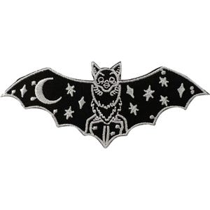 Könnte beinhalten: Ein schwarzer, bestickter Aufnäher in Fledermausform mit weißen Stickereien. Die Fledermaus zeigt eine zentrale Figur mit spitzen Ohren, einen zunehmenden Mond auf dem linken Flügel und verschiedene Sternformen, die über beide Flügel verteilt sind.