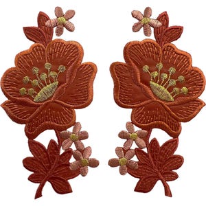 Pode incluir: Dois emblemas bordados simétricos com grandes flores cor de ferrugem com centros amarelo-dourado, rodeados por flores mais pequenas cor-de-rosa claro e folhas cor de ferrugem escuro. O bordado intrincado cria um design floral texturado.