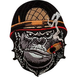 May include: An embroidered patch featuring a gorilla wearing a tan hat with a red band and a feather. The gorilla has grey fur, a stern expression, and is smoking a brown cigar with a thin trail of white smoke rising from the end of the cigar.